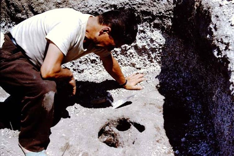 An old photograph, a man leans over a hole in the dirt