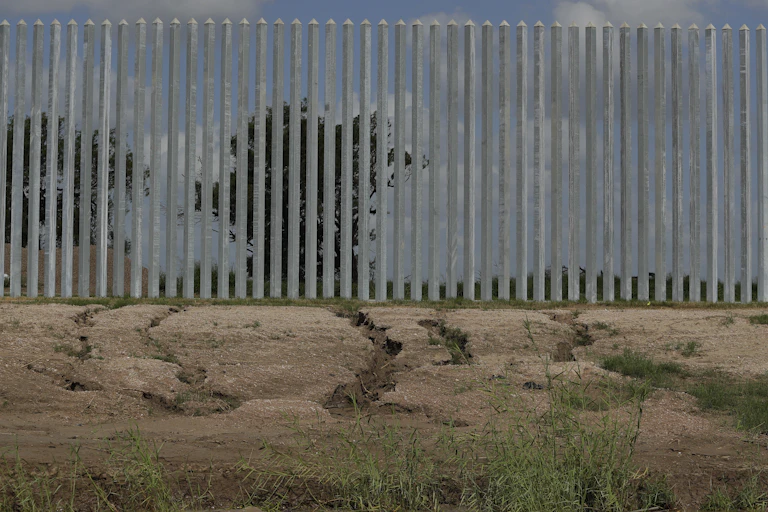 Cracks in the soil in front of a wall made of tall metal pillars.