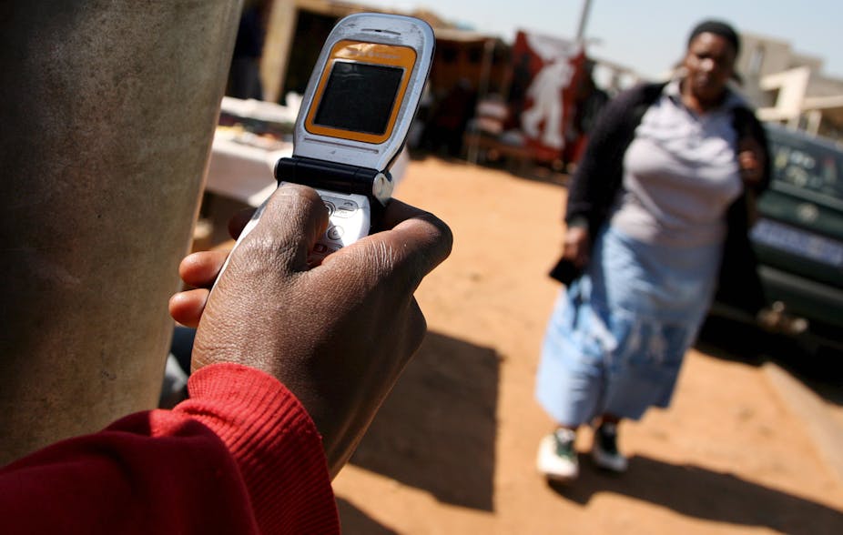 A man dials on a silver flip phone while a woman walks by on a dirt pavement in Soweto, South Africa