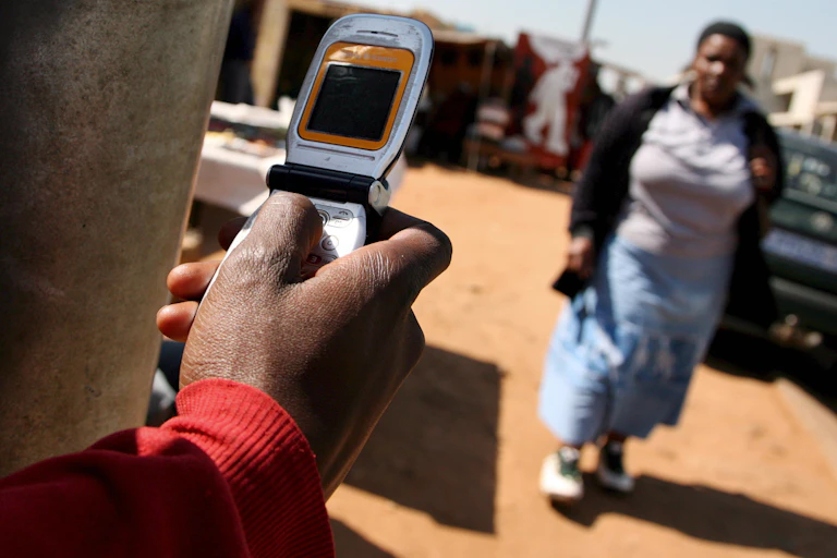 A man dials on a silver flip phone while a woman walks by on a dirt pavement in Soweto, South Africa