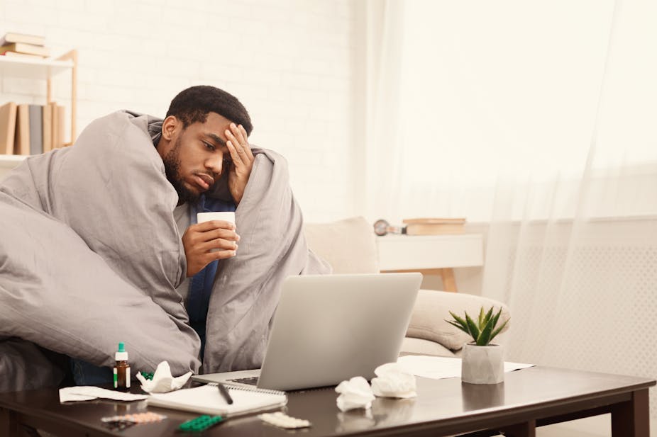 Man wrapped in duvet and holding head in front of laptop.