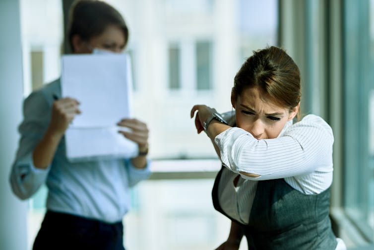 Young women coughs into her arm while colleague puts papers in front of her face.