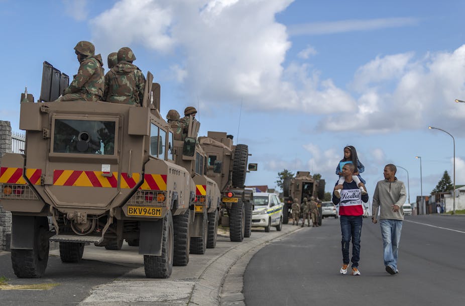 Two young man, one carrying a child on his shoulders, walk past a South African military convoy on a street in of Cape Town.