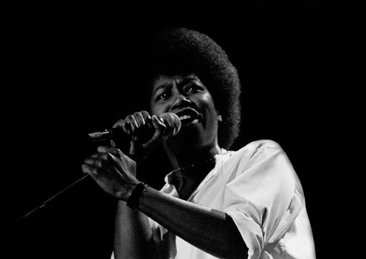 Black woman in white t-shirt singing into a microphone.