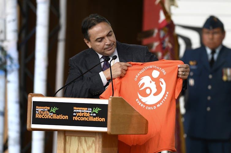 A man holds up an orange shirt at a podium.