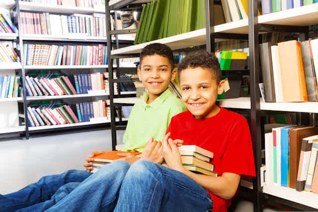 Two boys sitting on floor in a library, holding books and leaning against book shelf.