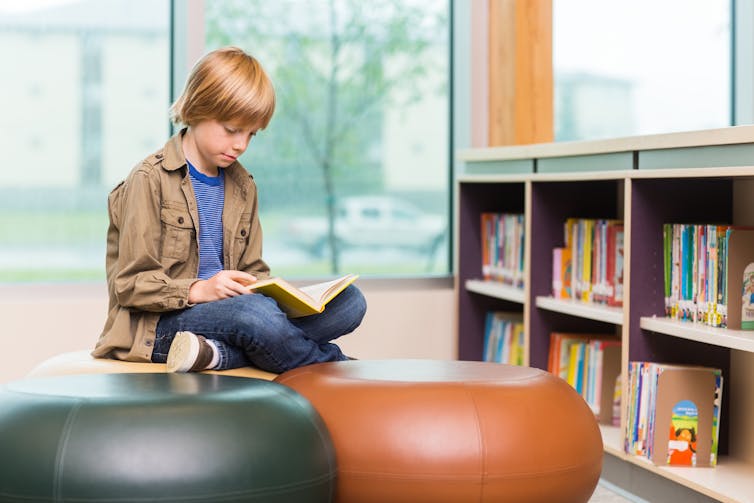 Boy sitting cross legged on round stool in library and reading.