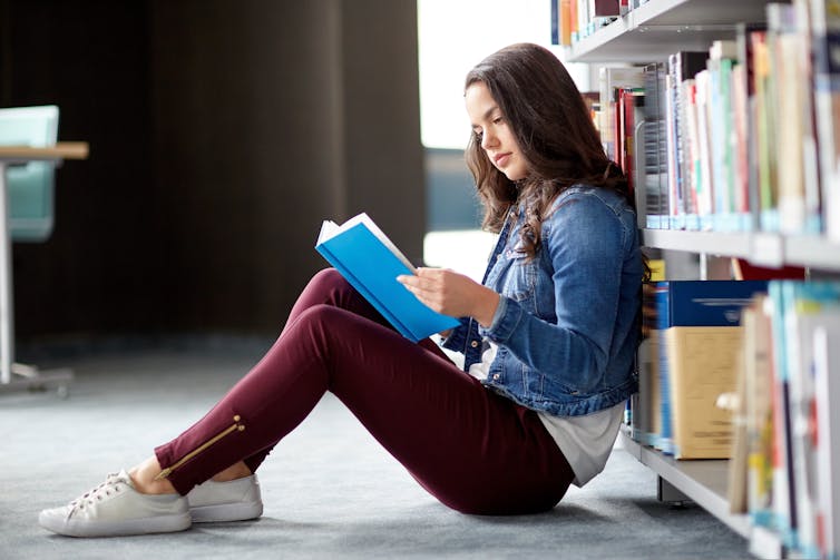 Girl reading in a library, leaning against book shelf.