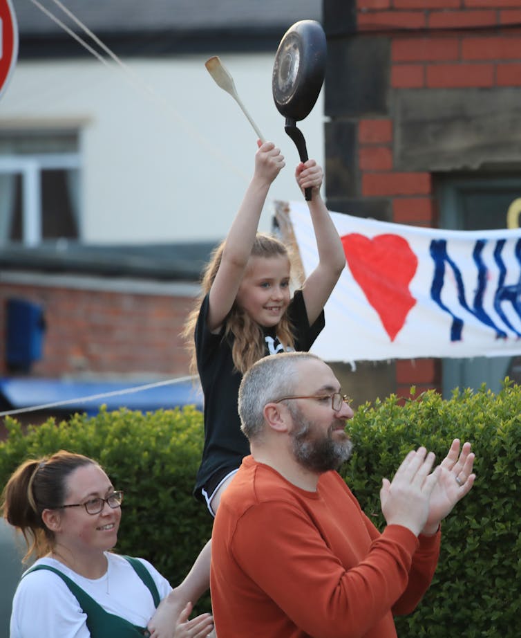 Two adults clap while little girl bangs frying pan
