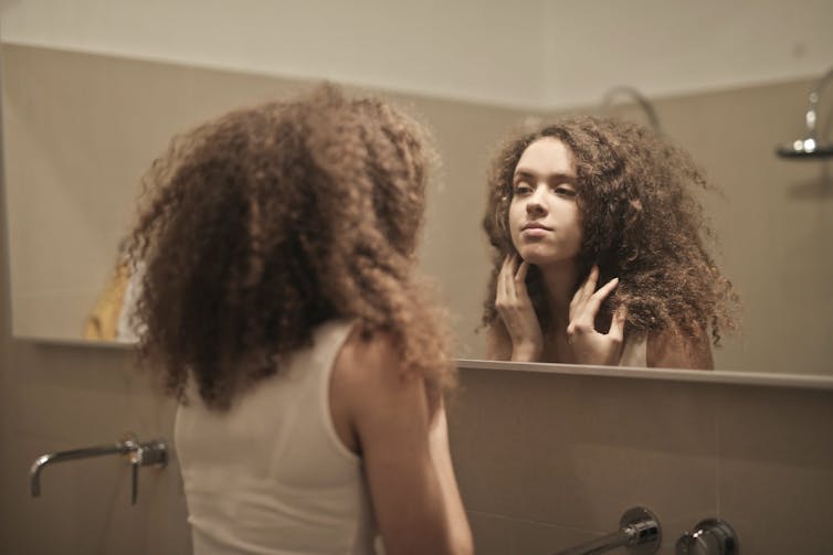 Woman in white tank top looking at herself in the mirror.