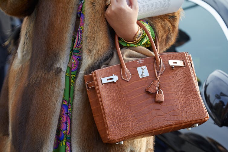 A woman wearing a fur coat holds a brown crocodile skin handbag.