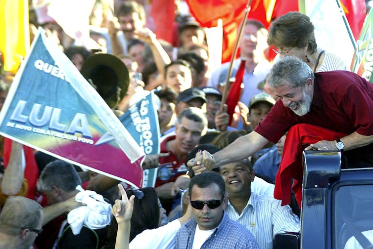 Lula da Silva on car greeting crowd of supporters holding flag with his name on it.