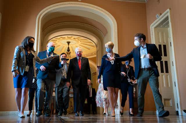 McConnell in a mask, walking through the Capitol surrounded by reporters recording him on cell phones 