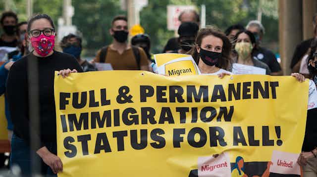 Women wearing masks carry a sign reading Full & Permanent Immigration Status for All