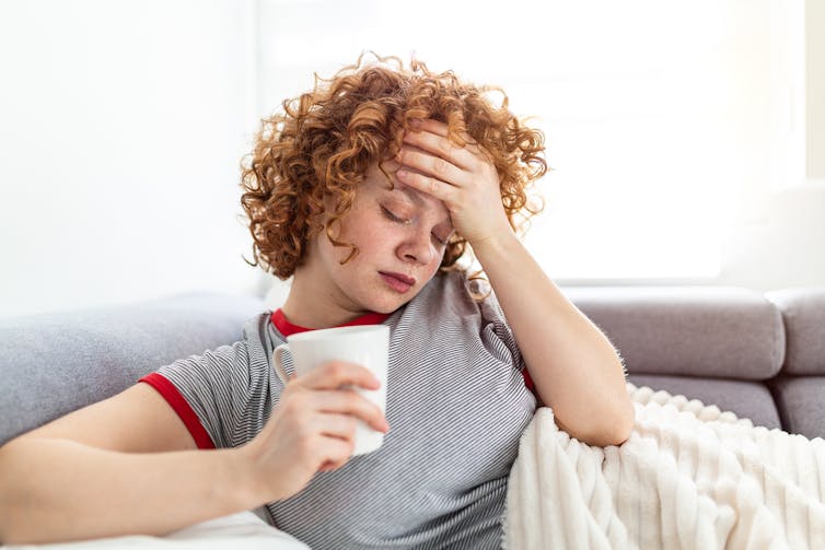 Young adult lying on sofa, clutching head, with cup in hand, sick