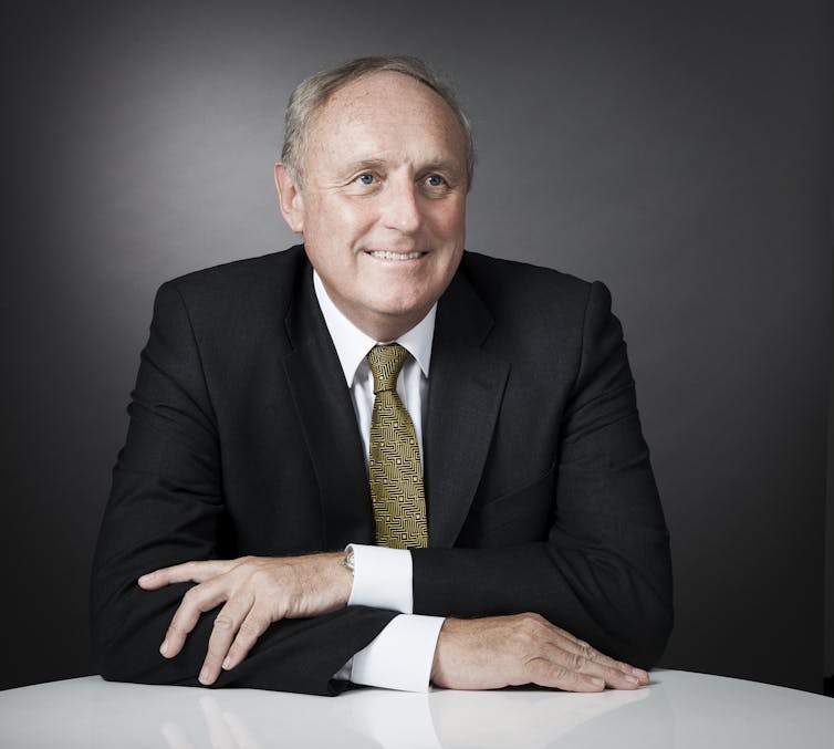 Head and shoulders shot of a middle-aged man in a suit and tie sitting at desk with his arms crossed.