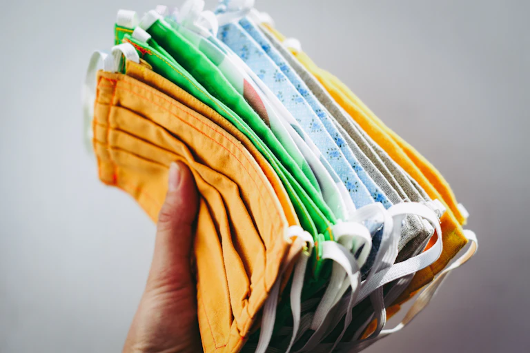 A hand holding a stack of cloth face masks in different colours: yellow, green and blue.