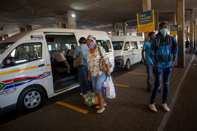 A woman wearing a COVID-19 mask, carrying a handbag and a platic bad with groceries, walks past a white minibus taxi at a rank.