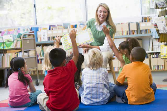 White teacher reading book to children sat on floor, hands up to answer question