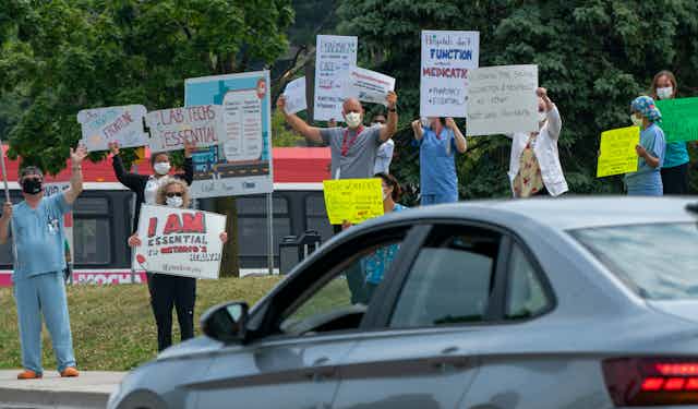 Health-care workers wearing masks and carrying signs wave at a passing car.