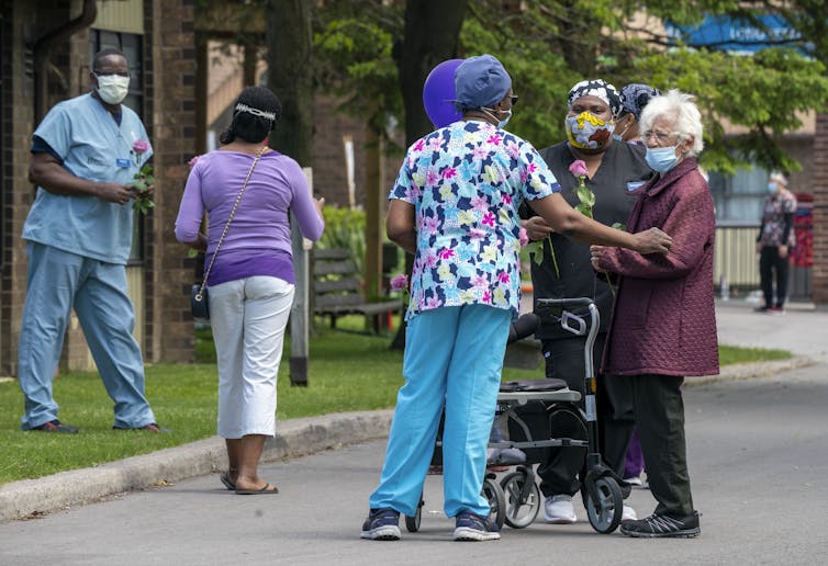 An elderly woman with a walker and wearing a mask chats with long-term workers wearing masks.