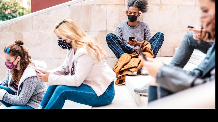Students sit on university steps looking at smartphones and wearing masks.