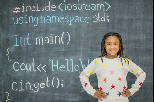 A Black girl with braids stands in front of a chalkboard with computer coding written in various colors