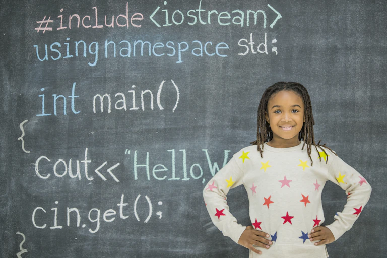 A Black girl with braids stands in front of a chalkboard with computer coding written in various colors