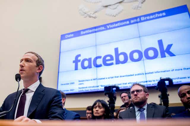 Mark Zuckerberg stands in front of a microphone with a screen showing the facebook logo behind him and the words Settlements, Violations and Breaches