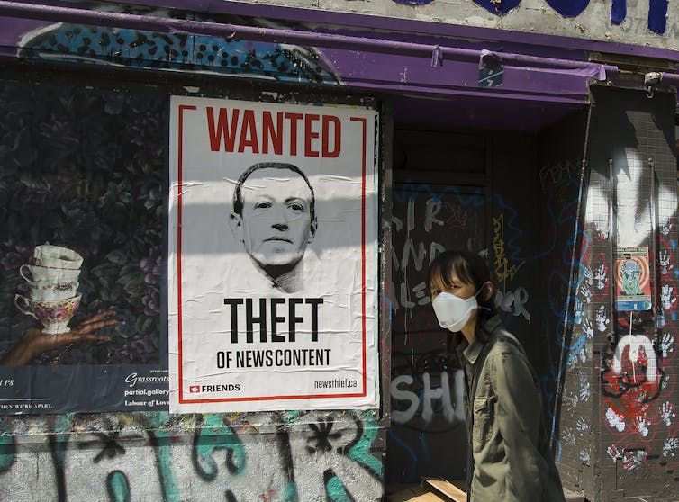 A woman wearing a surgical mask walks past a wanted poster of Mark Zuckerberg