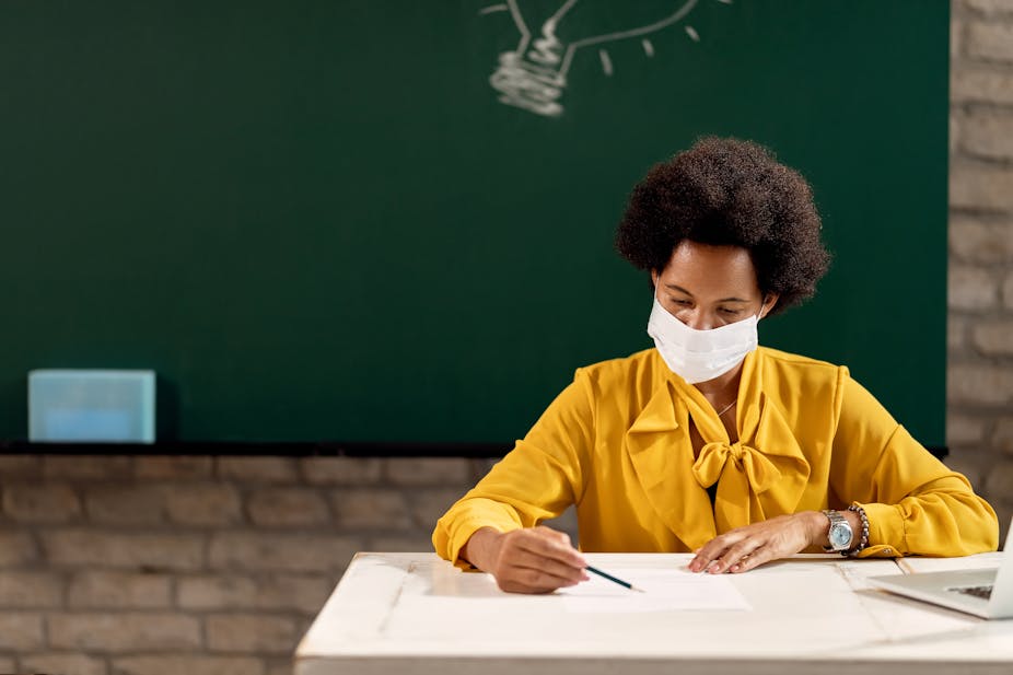 A teacher sitting on her desk with a face mask and a writing board behind her. 