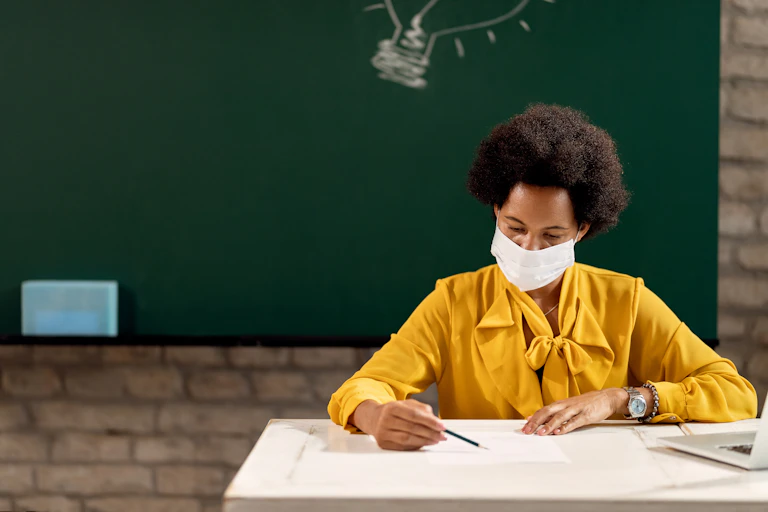 A teacher sitting on her desk with a face mask and a writing board behind her.