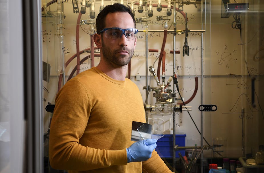 A man in a yellow shirt and blue latex gloves standing in front of a wall of equations holds a clear square of glass.