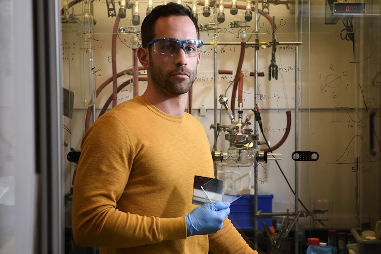 A man in a yellow shirt and blue latex gloves standing in front of a wall of equations holds a clear square of glass.