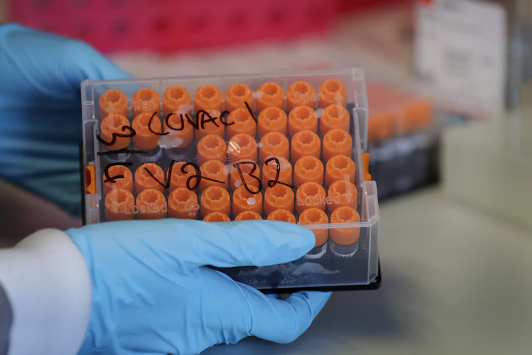 Hands in blue protective gloves handle a plastic case filled with rows of orange-topped vials.