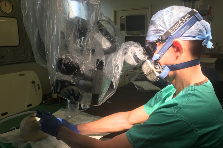 A surgeon in scrubs and a hairnet wears googles and a respirator mask and looks down a microscope while simulating an operation on a skull