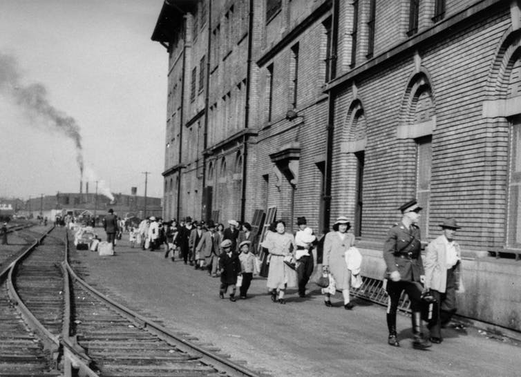 Black & white photo showing people walking past a building. Railroad tracks can be seen on the left of the image.