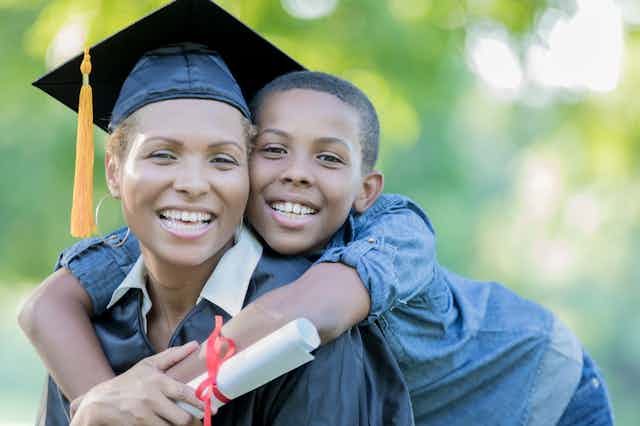A woman wearing a graduation cap is being hugged by her young son.