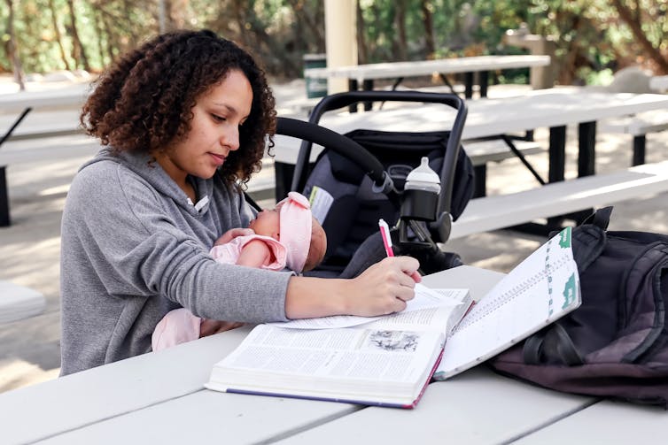 A mother holds a young baby while writing in a book at a table on a college campus.