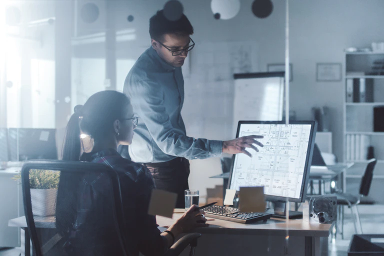 An older worker explains something on a computer screen to a young worker