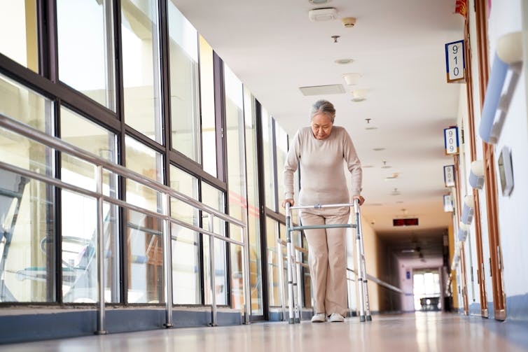 An elderly woman walks down the corridor of a nursing home using a frame.