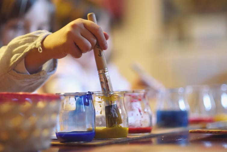 A kid's hand holding a paintbrush dipped in a jar of yellow paint.