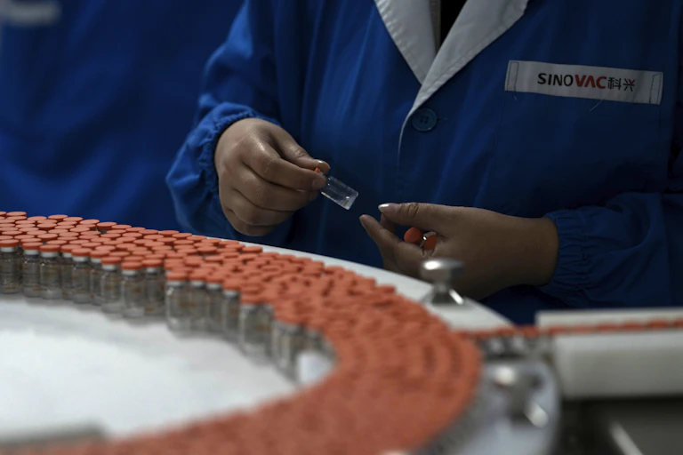Orange-capped vials on a conveyor belt. A worker holds several vials in their hands.