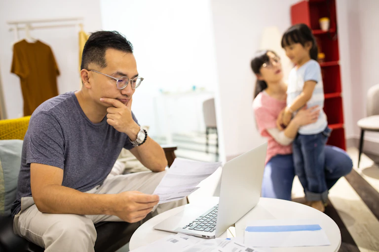 A man works on his computer while a woman in the background cares for a child.