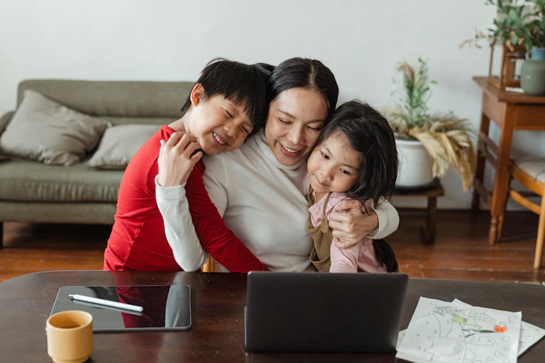 A woman sits at a table embracing her two children, a laptop in front of them.