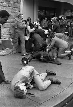 A chaotic scene of wounded people lying on a sidewalk while security officials scramble to protect other people.