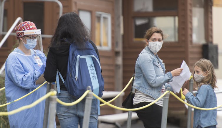 A health-care worker is seen wearing a face visor and other protective equipment as people wait in line to be tested for COVID-19.