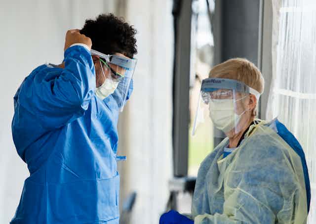 A health-care worker in personal protective equipment secures his visor as another health-care worker in full PPE stands near him.