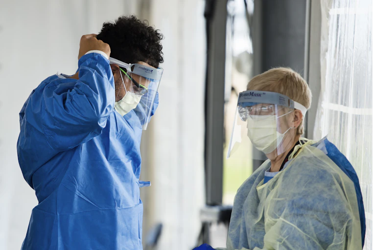 A health-care worker in personal protective equipment secures his visor as another health-care worker in full PPE stands near him.