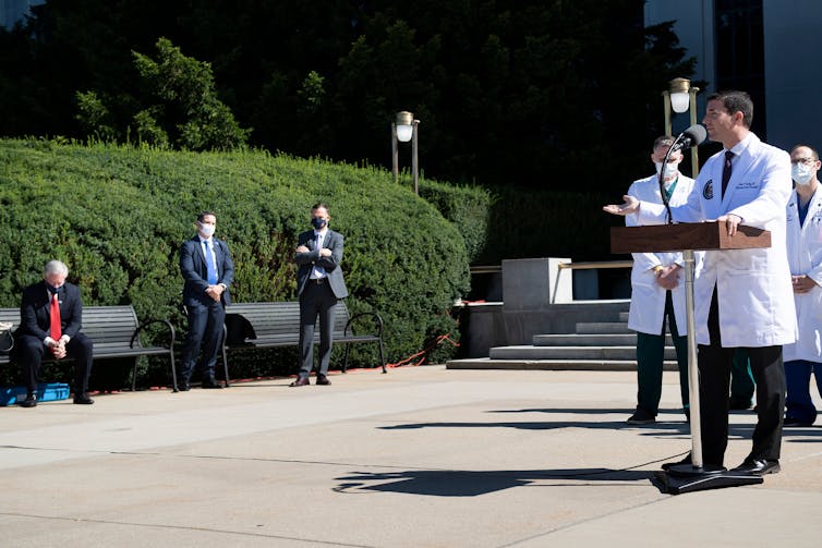 A man in a white doctor's jacket speaks to a few people wearing masks and staying at a distance.